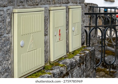 Outdoor electrical meter boxes mounted on a stone wall, beige power distribution cabinets with high voltage warning signs, technical infrastructure and utility enclosures on a granite facade near rail