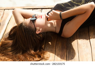 Outdoor fashion look of young woman wearing summer sunglasses,laying and laughing on the beach of sand,having sunbath.Wear summer bright outfit,vintage summer outfit colors.Hipster teenager girl