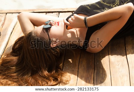 Outdoor fashion look of young woman wearing summer sunglasses,laying and laughing on the beach of sand,having sunbath.Wear summer bright outfit,vintage summer outfit colors.Hipster teenager girl