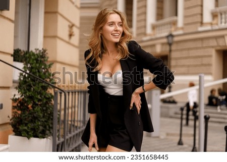 Outdoor fashion photo of beautiful blond woman dressed in   casual jacket, crop top and skirt.   Perfect smyle.   Walking in old european city. luxury live, trendy acsessorises.  