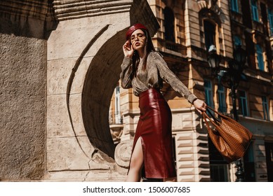 Outdoor fashion portrait of elegant, luxury woman wearing trendy leather  dark red skirt, beret, snakeskin print blouse, sunglasses, holding brown bag, walking in street of European city. Copy space