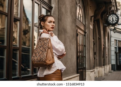 Outdoor fashion portrait of elegant, luxury woman wearing trendy white shirt, holding stylish beige, brown reptile, snakeskin, python print bag, handbag, posing in street. Copy, empty space for text 