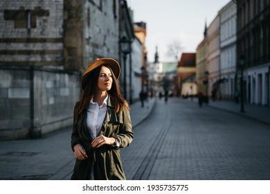 Outdoor fashion portrait of elegant, luxury woman wearing beige hat, sunglasses, trendy white shirt, in a green trench coat, walking in street. Copy, empty space for text. High quality photo