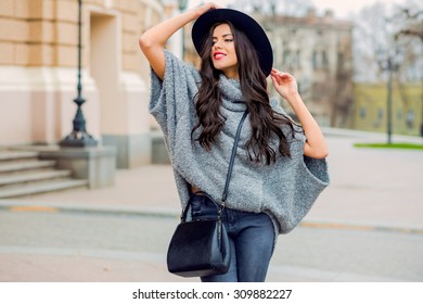 Outdoor fashion portrait of glamour sensual young stylish lady wearing trendy fall outfit , black hat , grey sweater and leather bag. Cold season.Red lips. Warm  clothes.