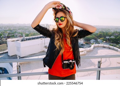 Outdoor fashion portrait of stylish photographer girl holding vintage retro camera, wearing bright swag hat, trendy sunglasses and leather jacket, amazing view of city from the roof. - Powered by Shutterstock