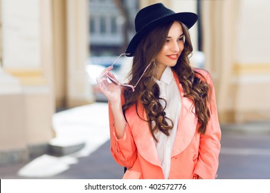 Outdoor  Fashion Portrait Of Stylish  Pretty Brunette  Woman In Night Spring Casual Outfit Walking In The City. Wearing White Blouse, Pink Jacket, Black Wool Hat. Long Wavy Hairstyle.