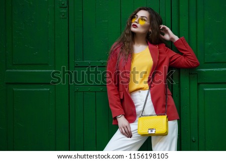 Outdoor fashion portrait of young beautiful woman wearing blazer, yellow sunglasses, blouse, white trousers, holding small quilted bag, posing near the green door. copy, empty space for text