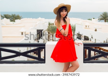  Outdoor fashion portrait of young pretty  happy woman in summer outfit posing in luxury resort on terrace  . Holiday vacation mood. Trendy hairstyle. Bright red dress. Flash tattoo. 