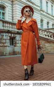 Outdoor Full Body Autumn Portrait Of Elegant Woman Wearing Long Orange Trench Coat, Hat, Sunglasses, Cowboy Ankle Boots, With Green Bag, Posing In Street Of European City