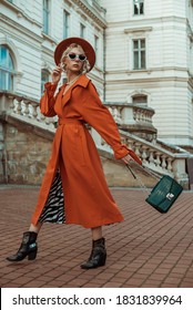 Outdoor Full Body Autumn Portrait Of Elegant Model Wearing Long Orange Trench Coat, Hat, Sunglasses, Cowboy Ankle Boots, With Green Croco Textured Bag, Posing In Street Of European City