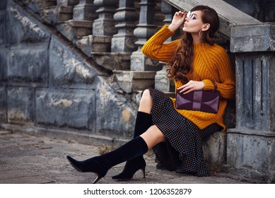 Outdoor full body fashion portrait of young beautiful woman wearing color sunglasses, stylish orange sweater, polka dot skirt, trendy sock boots, holding small purple bag, posing in street. Copy space