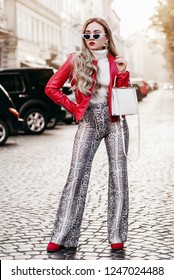 Outdoor full body fashion portrait of young fashionable lady wearing red biker jacket, sunglasses, turtleneck, trendy snake skin print trousers, holding white bag, posing in street of european city
