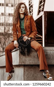 Outdoor Full Body Fashion Portrait Of Young Confident Fashonable Lady Wearing Brown Corduroy Blazer, Trousers, Green Turtleneck, Necklace, Cowboy Boots, Holding Suede Bag With Fringe, Posing In Street