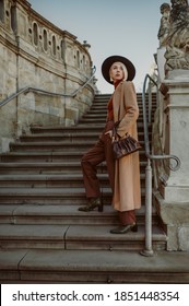 Outdoor full-length autumn fashion portrait of elegant, luxury woman wearing trendy long, midi beige, camel color coat, hat, classic trousers, trendy boots, holding brown leather pouch bag, handbag