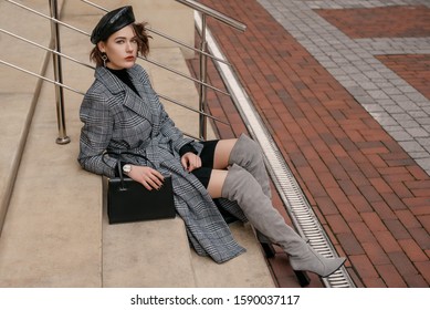 Outdoor full-length fashion portrait of  elegant woman wearing classic  checkered midi coat, gray suede high, over knee boots, beret, with black handbag, sitting, posing in street. Copy, empty space