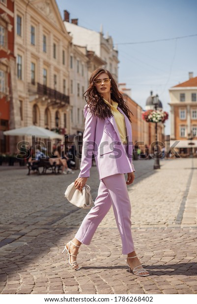 Outdoor\
full-length fashion portrait of elegant woman wearing lilac suit:\
blazer, trousers, strap sandals, holding trendy big white leather\
pouch handbag, walking in street of European\
city
