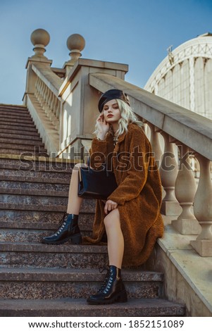 Outdoor full-length fashion portrait of young elegant woman wearing trendy brown faux fur coat, leather beret, lace up ankle boots, holding classic handbag, sitting, posing on stairs, in street