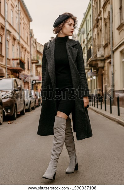 Outdoor\
full-length fashion portrait of young elegant woman wearing classic\
black coat, short turtleneck dress, gray suede high, over knee\
boots, beret, walking in street of European\
city