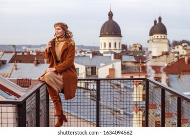 Outdoor full-length portrait of elegant fashionable happy smiling woman wearing trendy beige, brown outfit:  beret, dress, faux fur coat, high suede boots, posing in European city. Copy, empty space