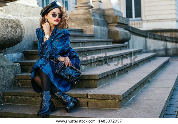 Outdoor full-length portrait of pretty curly lady
wearing trendy autumn checkered dress, leather beret, glasses,
wrist watch, black tights, boots, holding small bag, posing in
street. Copy space