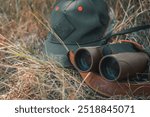 Outdoor gear featuring binoculars and a green cap lying in dry grass, symbolizing a nature hike, camping, or hunting scene, perfect for themes of exploration, adventure, or wildlife observation.