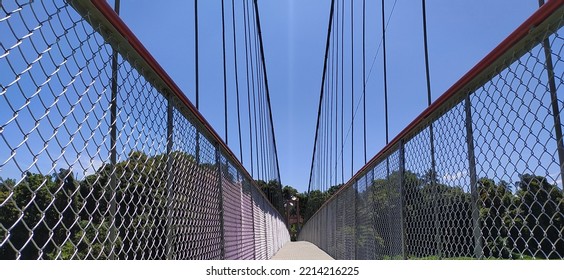 Outdoor Hanging Suspension Bridge Structure Made With Join Iorn Metal Cable Rope And Tower For Crossing Green Forest Lake Water. Empty And Quiet Forth Deck Walkway Or Path Close Up View Under Daylight