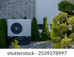 Outdoor heat pump unit set within well-kept garden of trimmed shrubs and stones, placed against brick wall with closed window shutter, forming energy-efficient residential setup. Modern heat pump. 