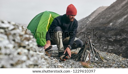 Outdoor horizontal image of hiker young male prepare brunches for bonfire in mountains. Traveler man with beard, sitting near to camping tent, making campfire after hiking. Travel, lifestyle concept.