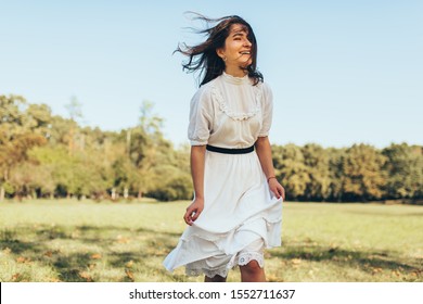 Outdoor image of beautiful happy woman with windy hair enjoying the warm weather, wearing white dress on nature background. Cheerful female smiling during walking in the park on sunny day.
