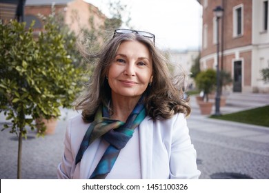 Outdoor image of gorgeous positive lady with charming smile and loose gray hair enjoying nice summer day, wearing white jacket, silk scarf and eyeglasses. Beauty, urban style and fashion concept