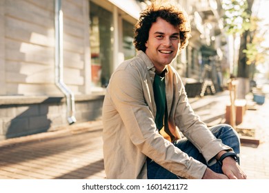 Outdoor image of happy smiling man with curly hair, posing for social advertisement sitting on the bench outdoors in the city street. Excited cheerful student male resting outside.