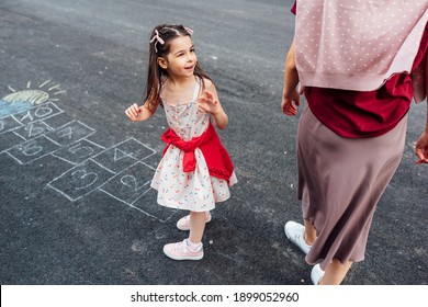 Outdoor Image Of A Little Girl Playing Hopscotch With Her Mother On Playground Outdoors. Child Plays With Her Mom Outside. Kid And Woman Plays Hopscotch Drawn On Pavement. Activities And Games Outside