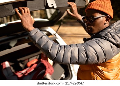 Outdoor Image Of Stylish African American Man Tourist In Spring Coat Sunglasses And Red Hat Closing Car Trunk After Packing And Collecting Things For Travel Or Going To Camping In Wild Nature