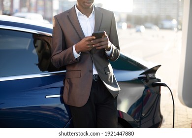 Outdoor image of of unrecognizable black man in formal business wear, leaning on his charging electric car and waiting for battery charge typing message on phone