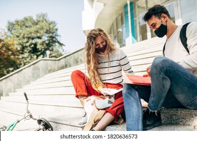 Outdoor Image Of A Young Woman And A Man Sitting On The Stairs Of The University For Team Work. Two Young Students In Masks Learning Together Sitting On The Stairs Of The College Campus Outside.