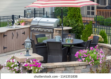Outdoor kitchen on a brick exterior patio with wicker dining chairs under an umbrella and colorful summer flowers on the circular wall