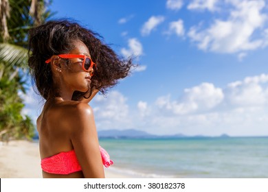 Outdoor Lifestyle Portrait Of Black Beautiful Teenage Girl In Pink Bra And Sunglasses. Young Hipster Woman Play With Her Hair. She Is Happy. Sunny Hot Summer Day At Tropical Ocean Beach With Palms.