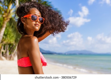 Outdoor Lifestyle Portrait Of Black Beautiful Teenage Girl In Pink Bra And Sunglasses. Young Hipster Woman Play With Her Hair. She Is Happy. Sunny Hot Summer Day At Tropical Sea Beach With Palms.