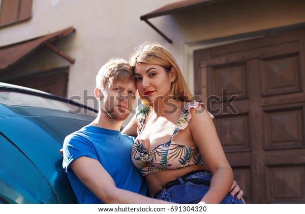 Outdoor lifestyle shot of romantic young\
Caucasian couple male and female posing outdoors, hugging, leaning\
on car and looking at camera. Cuddling, bonding, romance, dating\
and happiness concept