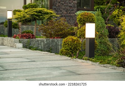 Outdoor Lights Installed Along Concrete Garden Path At The Residential House Backyard. Landscaped Flower Beds In The Background. Yard Illumination Theme.