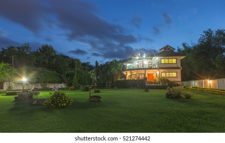 Outdoor Local Rural Private Isolated Beautiful Large Residential House At Night. Photo Taken Outside From The Front Garden Showing Decorative Yard Under A Dark Blue Cloudy Sky In A Summer Environment.