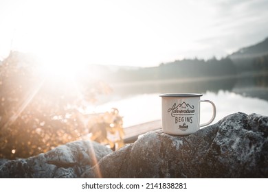 Outdoor Mug In Front Of A Lake And Forest With Blue Skies And Sun Flare In Background. The Adventure Begins Cup With Travel, Hiking And Camping Concept With Copy Space