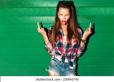 Outdoor night fashion urban portrait of young sexy pretty brunette spoiled crazy girl showing tongue and middle finger gesture on green background 