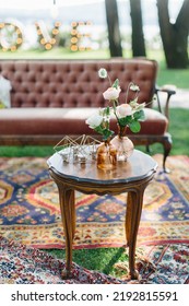 Outdoor Party Decor: In The Foreground Is A Vintage Wooden Table, Glass Vases With Roses Are In The Background, Geometric Golden Candlesticks With Candles. There Are Carpets On The Lawn, A Brown Sofa.