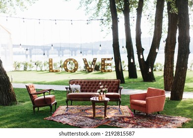 Outdoor Party Decor: A Vintage Wooden Table With Flowers In Vases In The Center, Sofas Next To It, Carpets On The Lawn. In The Background Are Glowing Letters 