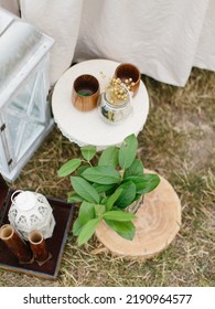 Outdoor Party Decor: A White Lantern On A Stand, A Twig With Green Leaves On A Wooden Stand, Wooden Cups On A White Table.