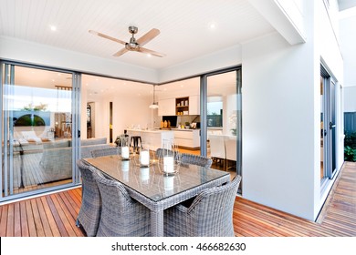 Outdoor Patio Area With Table Set Up Beside An Entrance To Inside Of A Modern House With A Kitchen, There Are Candles On The Table Under The Fan