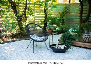 Outdoor patio furniture on pebbles in a front garden. Front veranda of house with black Acapulco armchair, coffee table and plants pots. Metal black fireplace bowl in garden on back yard patio. 
