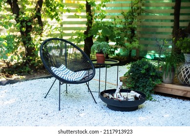 Outdoor patio furniture on pebbles in a front garden. Front veranda of house with two black Acapulco armchairs, coffee table and plants pots. Metal black fireplace bowl in garden on back yard patio. 
				