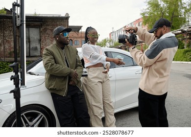 Outdoor photoshoot capturing two stylish individuals posing in front of white car while photographer adjusts camera. Urban scene with greenery and buildings in background - Powered by Shutterstock